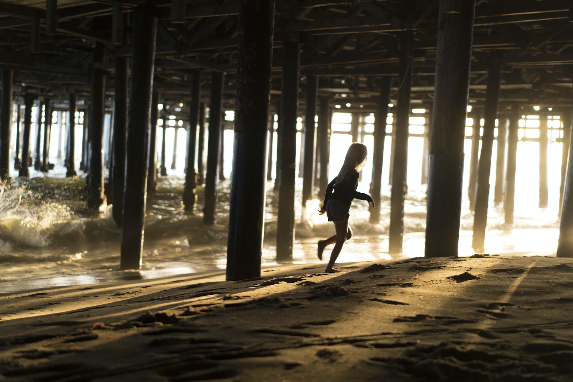 Silhouette of girl running from lapping waves underneath Santa Monica pier, California, USA