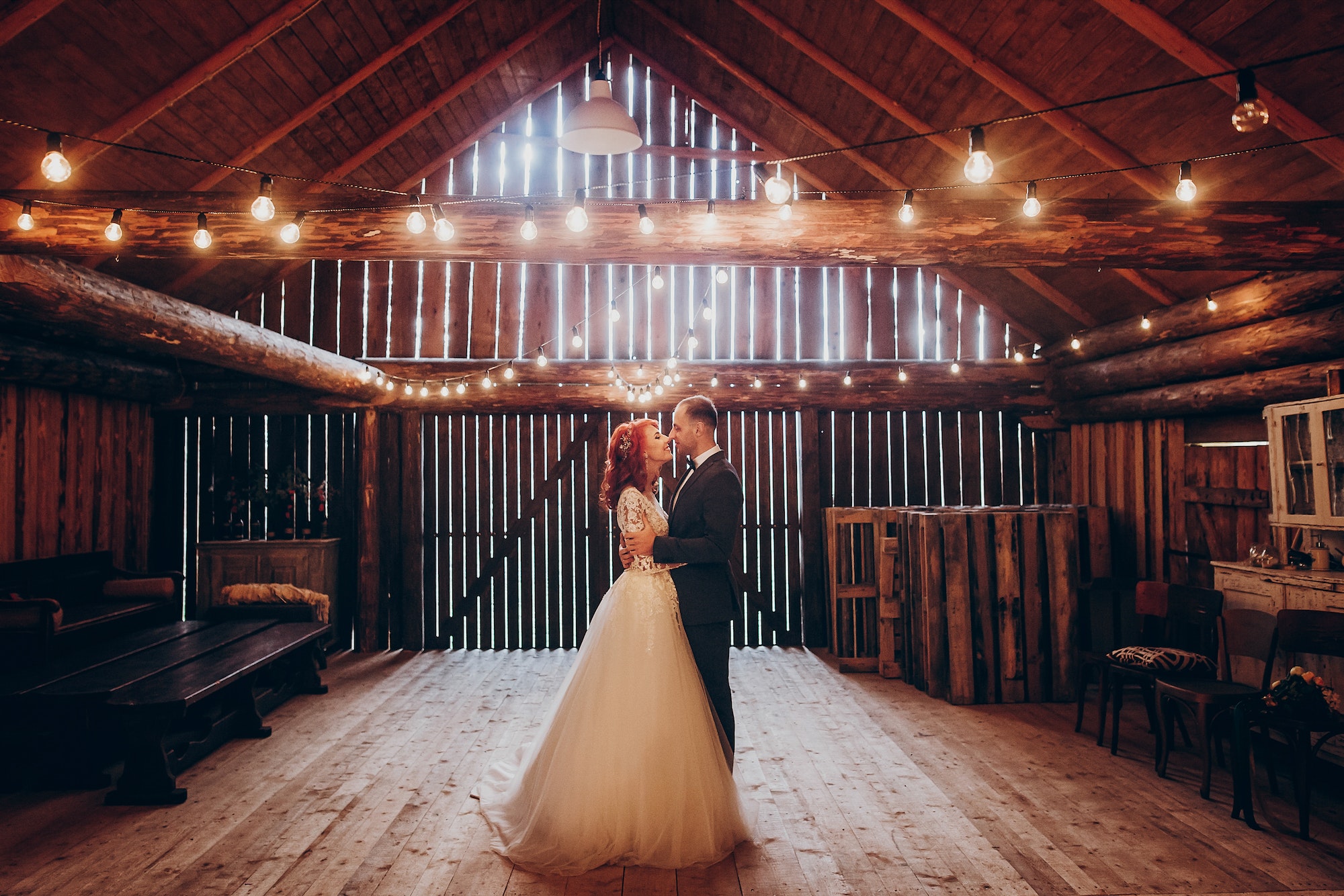 stylish groom and happy bride under retro bulbs lights in wooden barn